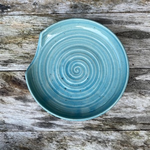 Könnte beinhalten: Eine runde, hellblaue Löffelablage aus Keramik mit einem eingeprägten Spiralmuster in der Mitte. Die Glasur hat eine glänzende Oberfläche mit kleinen, dunklen Sprenkeln, die über die Fläche verteilt sind, sowie eine Einkerbung für den Löffelstiel.