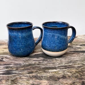 Könnte beinhalten: Zwei Keramikbecher mit einer tiefblauen Glasur und gesprenkelten Texturen. Die Becher haben einen abgerundeten Körper, einen geschwungenen Henkel und einen glatten, hellen Boden. Die Glasur verläuft von einem dunklen Blau zu einem helleren Farbton.