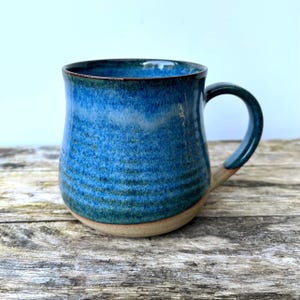 Könnte beinhalten: Eine handgefertigte Keramiktasse mit blauer Glasur und einem natürlichen, unglasierten Boden. Die Tasse hat einen gebogenen Henkel und eine strukturierte Oberfläche, ideal für Kaffee oder Tee. Die Abmessungen der Tasse sind nicht sichtbar.