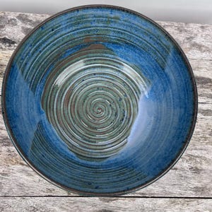 Könnte beinhalten: Eine runde Keramikschale mit einer tiefblauen Glasur und sichtbaren konzentrischen Spiralrillen, die in die Innenfläche eingearbeitet sind. Die Mitte der Schale zeigt einen gedämpften Grün- und Braunton, während der Rand dunkelbraun ist.