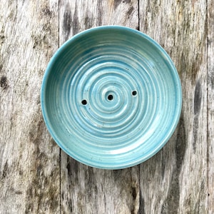 Könnte beinhalten: Eine runde, hellblaue Keramik-Seifenschale mit einem eingeprägten Spiralmuster auf der Innenfläche. Die Schale hat eine gesprenkelte Oberfläche und verfügt über drei kleine Abflusslöcher in der Nähe der Mitte des Spiraldesigns.
