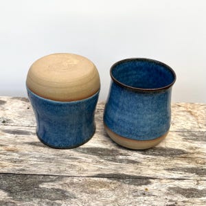 Könnte beinhalten: Zwei handgefertigte Keramikteile. Eines ist eine abgerundete Form mit einem beigefarbenen Oberteil und einem blauen Sockel. Das andere ist eine blaue Tasse mit einem beigefarbenen Sockel und einem dunkelbraunen Rand. Beide stehen auf einer Holzoberfläche.