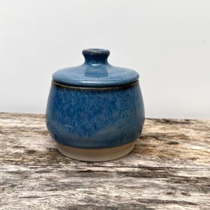 Könnte beinhalten: Ein Keramikgefäß mit Deckel, das eine glänzende blaue Glasur mit dunkleren blauen gesprenkelten Akzenten aufweist. Das Gefäß hat einen abgerundeten Körper, einen kleinen Knauf auf dem Deckel und einen kontrastierenden beigen Boden.