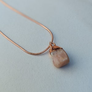 May include: A delicate rose gold chain necklace with a raw, light pink sunstone pendant.