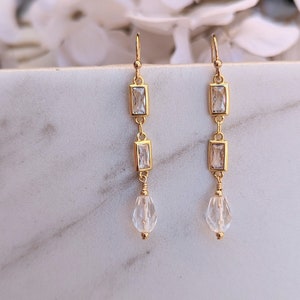 May include: Gold dangle earrings with three clear rectangular stones and a teardrop-shaped crystal at the bottom.