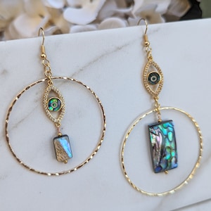 Könnte beinhalten: Goldene Creolen mit einem baumelnden bösen Auge und einem rechteckigen Abalone-Muschel-Anhänger.