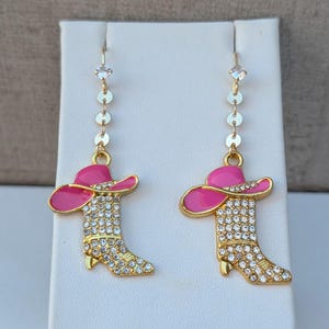 Könnte beinhalten: Ein Paar goldfarbene Ohrhänger mit einem pinkfarbenen Emaille-Cowboyhut, der auf einem mit Strasssteinen besetzten Cowboystiefel sitzt. Die Ohrringe hängen an einer goldfarbenen Kette, die mit kleinen klaren Kristallen verziert ist.