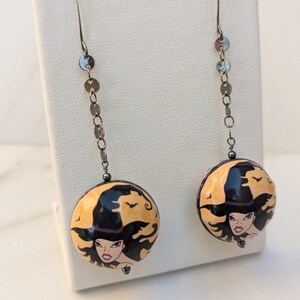 May include: A pair of dangle earrings with a witch face charm. The charms are round and feature a witch with a black hat and long black hair. The earrings are silver and have a chain of small silver circles.
