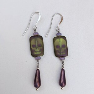 May include: A pair of silver skull earrings with green and purple glass beads. The skull is a translucent green with a purple outline. The beads are a deep purple.