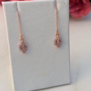 May include: Rose gold cactus earrings with clear crystal accents. The earrings hang from delicate chains and are displayed on a white surface. The background features pink roses, adding a touch of color to the image.