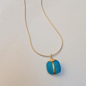 May include: A gold-toned chain necklace featuring a square-shaped, bright turquoise stone pendant. The stone is secured with gold-colored wire wrapping that extends from the top bail down the center of the stone, creating a secure, decorative setting.