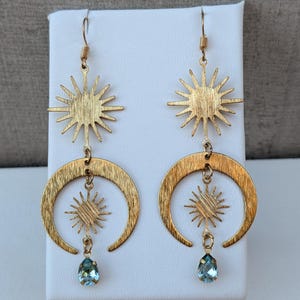 May include: A pair of gold-toned dangle earrings featuring a sunburst charm at the top, a crescent moon shape in the center, a smaller sunburst charm suspended within the moon, and a single light blue teardrop-shaped crystal at the bottom.