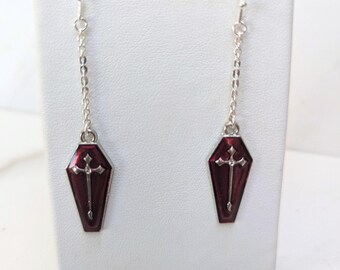 May include: A pair of dangle earrings featuring silver-toned metal chains attached to coffin-shaped pendants. Each pendant is filled with deep red enamel and displays a raised silver-toned cross centered on the surface of the coffin shape.