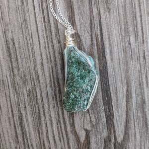 May include: A green gemstone pendant wrapped in silver wire, hanging from a silver chain. The pendant is a rough, irregular shape and has a dark green colour with flecks of lighter green.