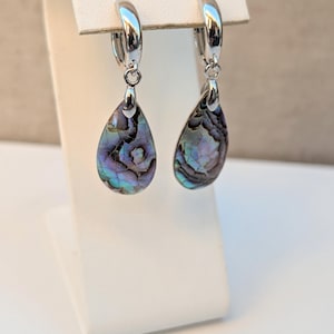 May include: Silver-tone hoop earrings with teardrop pendants. The pendants are made from iridescent abalone shells, displaying swirling patterns of blue, green, and purple. The earrings are presented on a white stand.