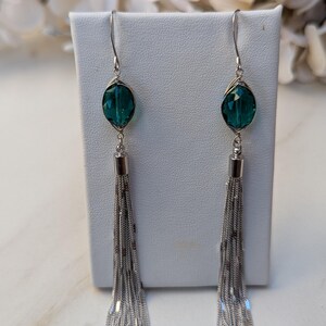 May include: Elegant dangle earrings featuring teal-colored faceted glass beads and long, silver-tone chain tassels. The earrings have a simple hook design and are displayed on a white stand, showcasing their delicate details.