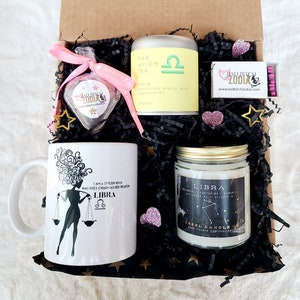 May include: A gift box filled with Libra themed items, including a mug, candle, tea, and chocolate. The mug features a silhouette of a woman with a Libra symbol and the text "I am a stylish bitch who uses charm as her weapon Libra". The candle is white with a gold lid and the text "Libra" and "Balance, Fascinating, Romantic September 23 - October 22". The tea is in a green tin with a Libra symbol and the text "Par Avion Tea Libra micccan blend with peppermint". The chocolate is in a heart-shaped box with a Libra symbol and the text "Bad Bitch Zodiac".