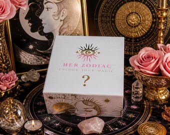 Op de afbeelding: Een witte rechthoekige doos met een gouden oogpictogram, de tekst HER ZODIAC UNLOCK YOUR MAGIC en een gouden vraagteken. De doos is versierd met gouden hemelse illustraties van zonnen en manen, omringd door roze rozen en kristallen.