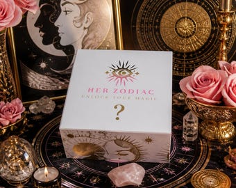 Op de afbeelding: Een witte rechthoekige doos met een gouden oogpictogram, de tekst HER ZODIAC UNLOCK YOUR MAGIC en een gouden vraagteken. De doos is versierd met gouden hemelse illustraties van zonnen en manen, omringd door roze rozen en kristallen.