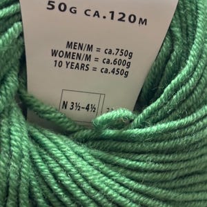 May include: A close-up shot of a ball of green yarn with a label attached. The label shows a barcode, the yarn's weight (50g), and its approximate length (120m). The label also provides yarn usage estimates for different age groups.