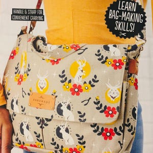 Puede incluir: El Suzie Satchel, un bolso de tela beige con cierre de solapa, presenta un patrón repetido de conejos blancos, círculos amarillos y flores rojas. El bolso tiene un asa y una correa para llevarlo, y el texto "Learn Bag-Making Skills!" es visible.