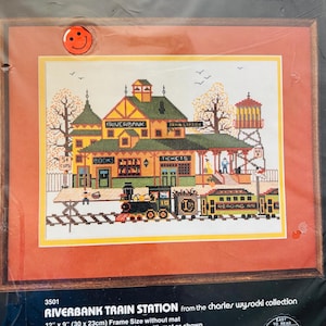 Puede incluir: Un kit de punto de cruz contado que representa la estación de tren de Riverbank. El diseño incluye un tren, una estación y una torre de agua, con el texto "Riverbank Train Station" y "Tickets". El tamaño del marco es de 30 x 23 cm sin paspartú.