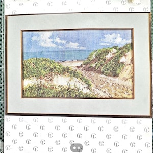 May include: A rectangular cross-stitch embroidery depicting a coastal landscape with green sand dunes, a sandy path, and a blue ocean under a cloudy sky. The artwork is framed in a thin brown wooden frame with a wide, light grey matting border.