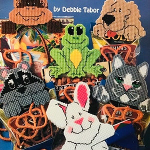 Op de afbeelding: Een set van zes plastic canvas snackbag clips met verschillende dierenontwerpen. De clips zijn in de vorm van een aap, hond, kikker, wasbeer, konijn en kat. De clips zijn omringd door zakken met pretzels.