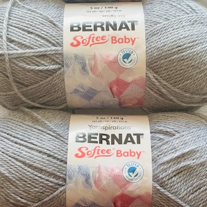 Könnte beinhalten: Zwei Knäuel hellgraues Bernat Softee Baby Garn, jeweils mit einem Papieretikett mit der Aufschrift 5 oz / 140 g und 362 yds / 331 m. Das Etikett zeigt den Markennamen, den Produktnamen und ein kreisförmiges Logo für geprüfte Schadstofffreiheit.