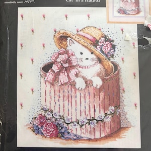 May include: Cross-stitch kit titled "Cat in a Hatbox" featuring a white cat wearing a straw hat and sitting in a pink hatbox. The kit includes Aida fabric, floss, needle, graph, and instructions. Finished size is 31.8 cm x 35.6 cm.