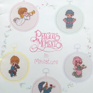 Puede incluir: Una hoja de patrones de punto de cruz con personajes de Precious Moments en miniatura. Los diseños incluyen un ángel tocando la trompeta, un niño sosteniendo un oso de peluche y otras figuras. Se muestra el texto "Precious Moments in Miniature".