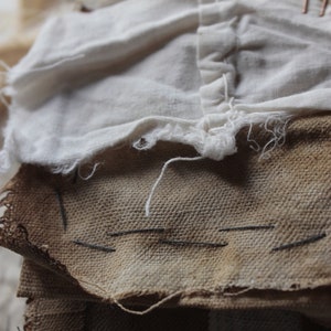 Puede incluir: Primer plano de una pila de recortes de tela, incluyendo una pieza blanca con un borde deshilachado y una pieza marr&oacute;n con costuras visibles. La tela es texturizada y tiene un aspecto r&uacute;stico.