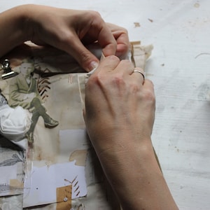 Puede incluir: Una persona est&aacute; trabajando en un diario hecho a mano con una flor blanca, una imagen vintage y un clip. El diario est&aacute; sobre una superficie blanca con un carrete de hilo negro y un trozo de tela blanca.