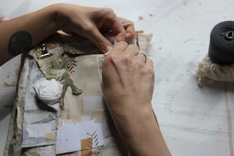 Puede incluir: Una persona est&aacute; trabajando en un diario hecho a mano con una flor blanca, una imagen vintage y un clip. El diario est&aacute; sobre una superficie blanca con un carrete de hilo negro y un trozo de tela blanca.