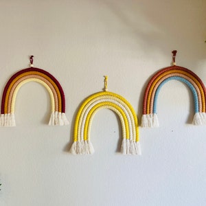 May include: Three colorful macrame rainbows hanging on a white wall. The rainbows are made with different colored yarn, including red, orange, yellow, blue, and white.