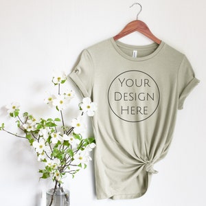 May include: A sage green t-shirt with rolled up sleeves hangs on a wooden hanger. The shirt has a circle on the front with the text "YOUR DESIGN HERE" inside.