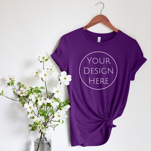 Könnte beinhalten: Lilafarbenes T-Shirt mit einem weißen Kreis auf der Vorderseite, auf dem "YOUR DESIGN HERE" steht. Das Shirt hängt an einem Holzbügel an einer weißen Wand. Links neben dem Shirt steht eine Vase mit weißen Blumen.