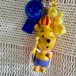 May include: A keychain featuring a yellow bear figure wearing a red and yellow crown, a white cape, and purple shorts, holding a small gold scepter. Attached are a blue plastic whistle, a purple ball chain, and a translucent yellow bear charm.