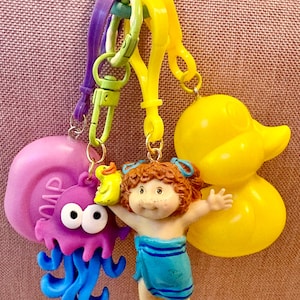 May include: A collection of colorful keychains featuring a purple soap, a purple jellyfish, a yellow rubber duck, and a doll wearing a blue towel. The keychains are attached to a light blue and yellow clasp.