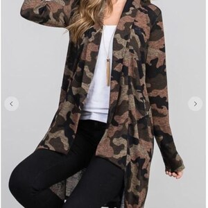 A blonde model wearing a hat, a brown camouflage long cardigan over a white top and black pants, and sitting on a stool.