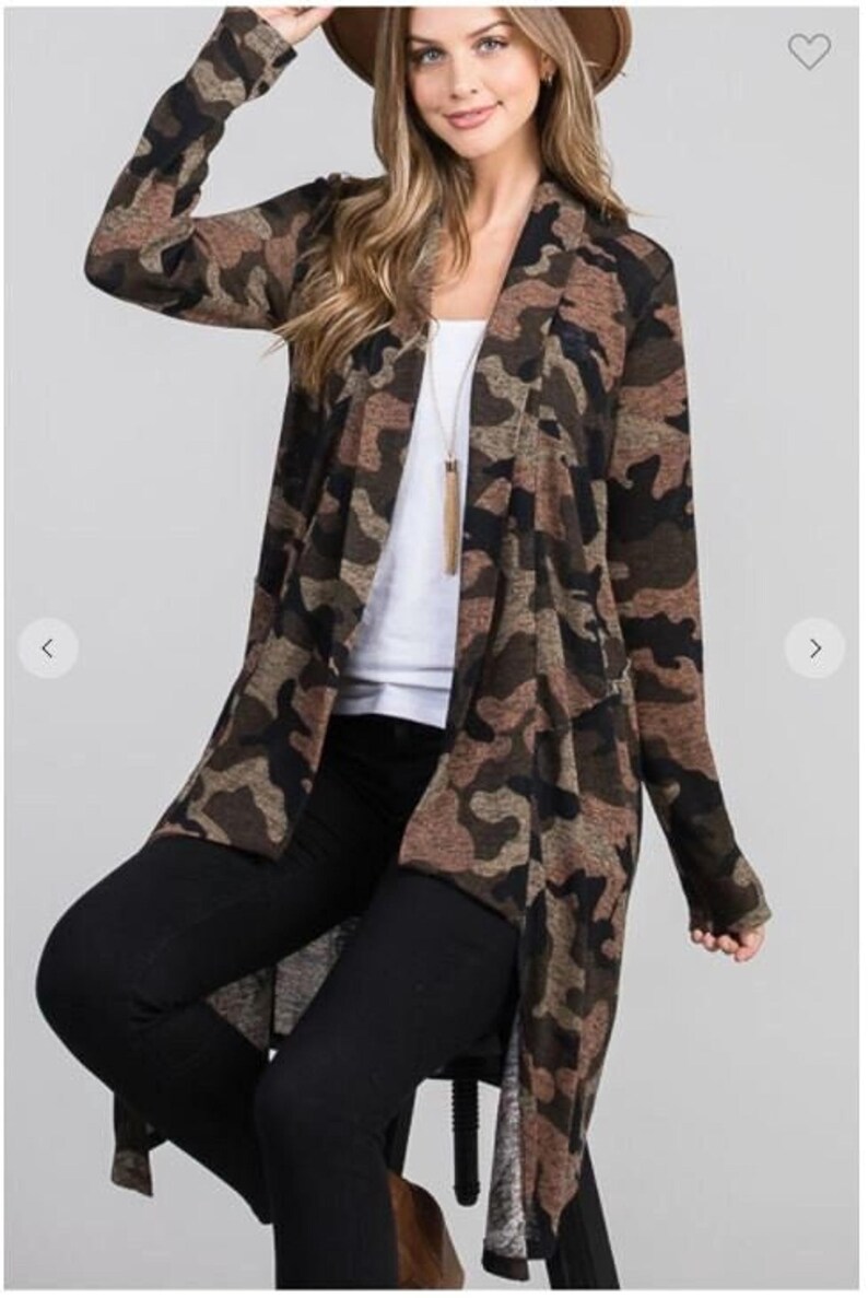 A blonde model wearing a hat, a brown camouflage long cardigan over a white top and black pants, and sitting on a stool.