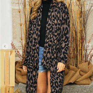 A blonde model wearing round sunglasses and a long brown animal print cardigan over a black top and denim shorts against a textured backdrop.