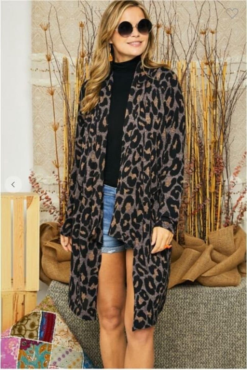 A blonde model wearing round sunglasses and a long brown animal print cardigan over a black top and denim shorts against a textured backdrop.