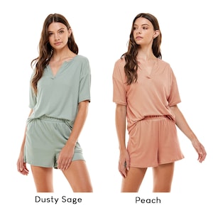 May include: Two women wearing matching ribbed short-sleeved tops and shorts. The woman on the left is wearing a dusty sage green set, and the woman on the right is wearing a peach set. Both tops have a V-neckline.