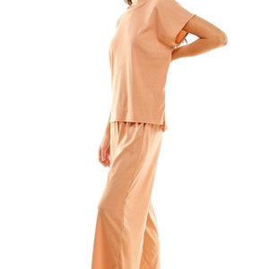 May include: A peach-coloured two-piece lounge set with a short-sleeved top and wide-leg trousers. The model is wearing the set and white sandals.