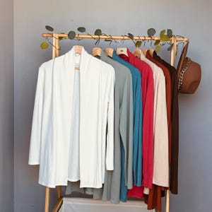 May include: A clothing rack displays a variety of long-sleeved cardigans in shades of white, gray, blue, red, beige, and brown. A brown hat with a chain detail hangs on the right side. The cardigans are made of a soft material.