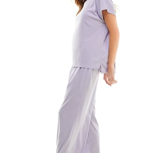 May include: A lavender-coloured two-piece pyjama set with a short-sleeved top and wide-leg trousers. The model is wearing white trainers.