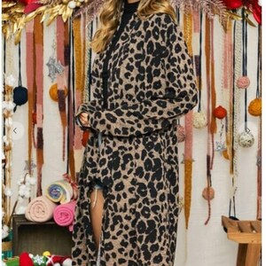 A blonde model wearing round sunglasses, a black turtleneck, and a long brown and black animal print cardigan in front of a rustic fabric backdrop.