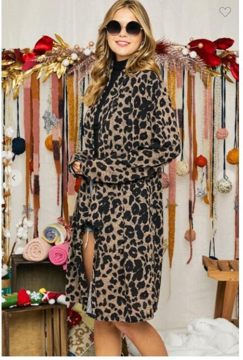 A blonde model wearing round sunglasses, a black turtleneck, and a long brown and black animal print cardigan in front of a rustic fabric backdrop.