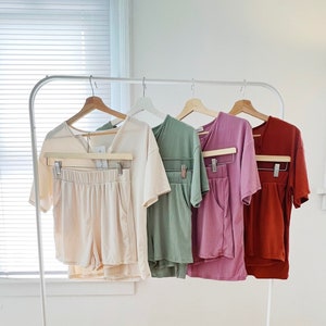 May include: Four sets of matching pajamas, each with a short-sleeved top and shorts. The pajamas are displayed on hangers and are in the colors beige, green, purple, and rust.